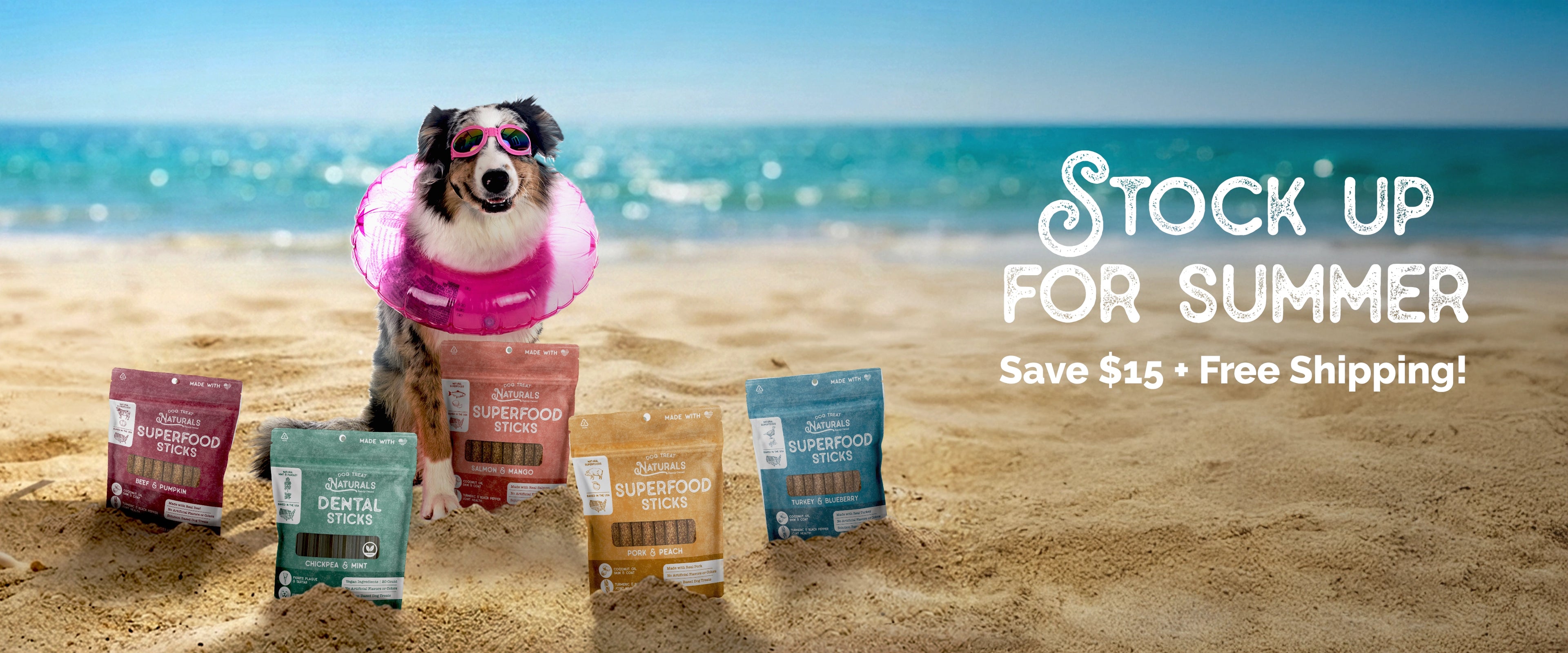 A dog wearing a pink swim ring stands on a sandy beach with the ocean in the background, surrounded by several bags of dog treats. Large text on the right reads 'Stock Up for Summer Save $15 + Free Shipping!'.